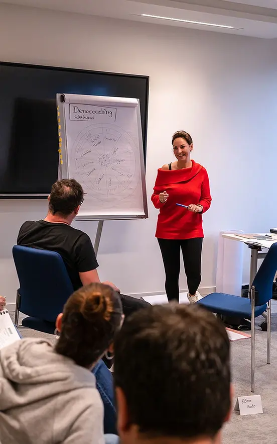 Eine Frau im roten Pullover hält einen Vortrag mit einem Clipboard vor einer kleinen Gruppe Menschen mit