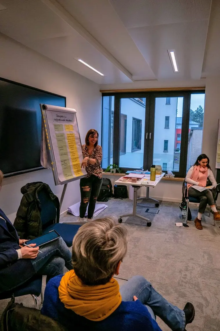 Eine Frau in zerrissenen Jeans und einer geblümten Bluse hält einen Vortrag vor einem Clipboard