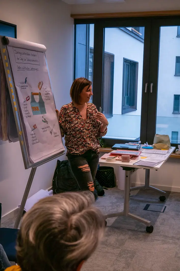 Eine Frau in zerrissenen Jeans und einer geblümten Bluse hält einen Vortrag mit einem Clipboard vor einer kleinen Gruppe Menschen mit