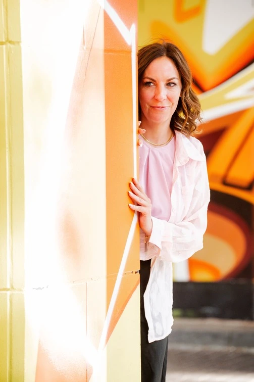 Ramona Leis - Pädagogin für Mindset-Entwicklung - Nahaufnahme von einer Frau mit welligem, braunen Haar, sie schaut hinter einer Wand hervor