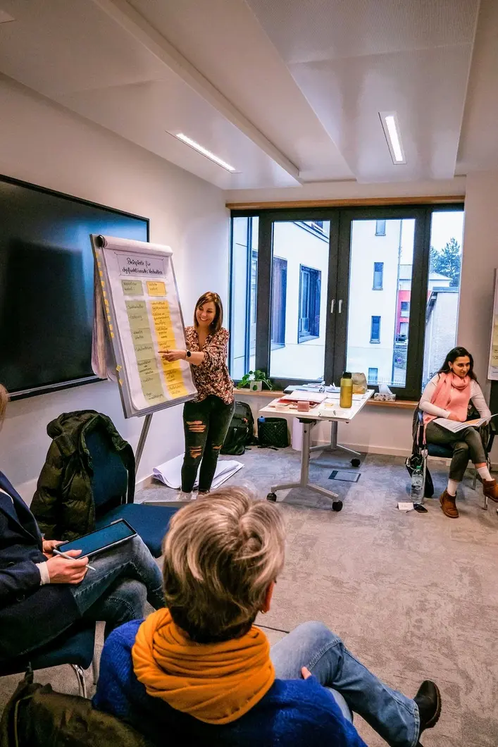 Eine Frau in zerrissenen Jeans und einer geblümten Bluse hält einen Vortrag und zeigt mit dem Finger auf ein Clipboard, Ausbildung zum Mindset-Pädagoge